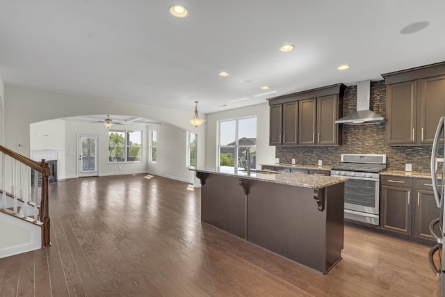 kitchen with light stone counters, stainless steel range, a breakfast bar, ceiling fan with notable chandelier, and wall chimney range hood