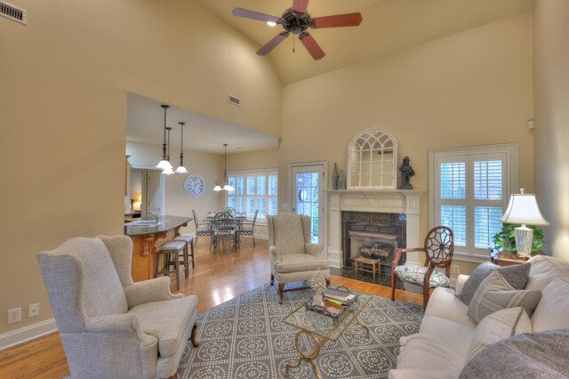 living room with ceiling fan with notable chandelier, light wood-type flooring, high vaulted ceiling, and a tiled fireplace