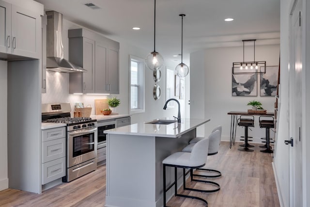 kitchen with wall chimney range hood, sink, hanging light fixtures, a kitchen island with sink, and gas range