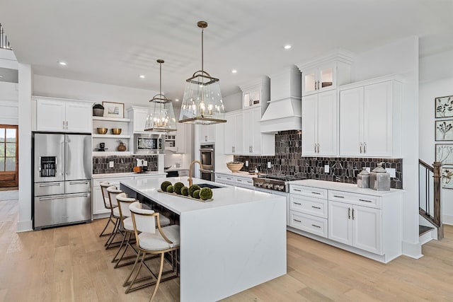 kitchen featuring appliances with stainless steel finishes, white cabinetry, custom range hood, and an island with sink