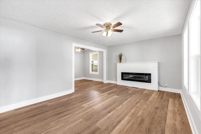 unfurnished living room featuring a ceiling fan, a textured ceiling, wood finished floors, a glass covered fireplace, and baseboards