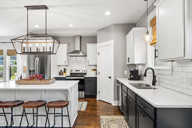 kitchen with sink, stainless steel appliances, wall chimney range hood, pendant lighting, and white cabinets