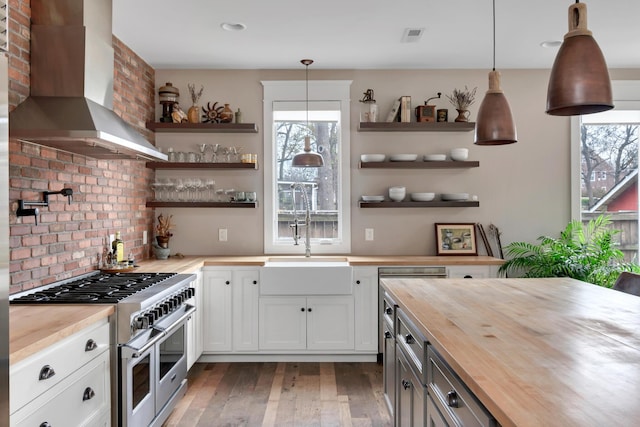 kitchen with range with two ovens, wall chimney range hood, butcher block countertops, decorative light fixtures, and white cabinetry