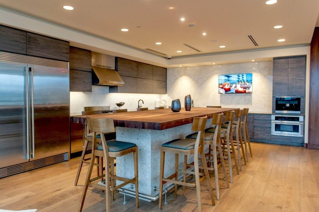 kitchen with butcher block counters, wall chimney range hood, a breakfast bar area, appliances with stainless steel finishes, and light wood-type flooring