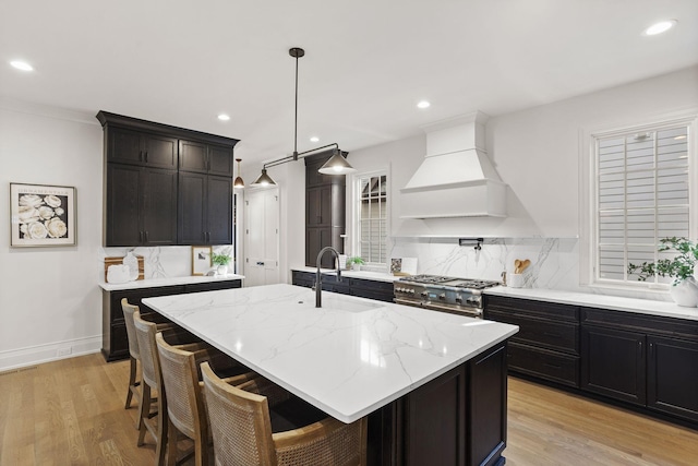 kitchen featuring sink, premium range hood, high end stove, a kitchen island with sink, and light wood-type flooring