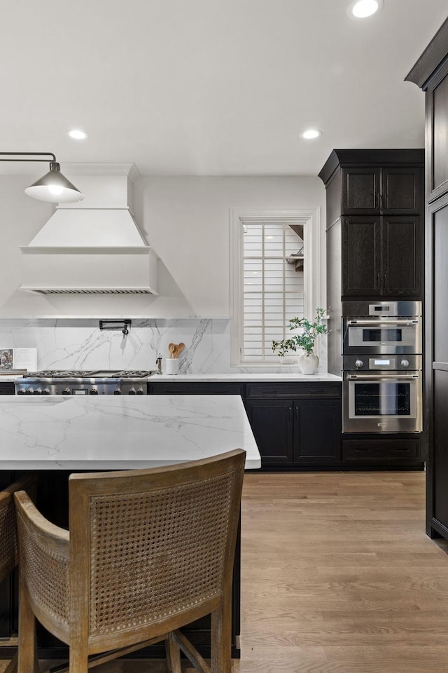 kitchen with custom exhaust hood, backsplash, light hardwood / wood-style floors, light stone counters, and stainless steel double oven