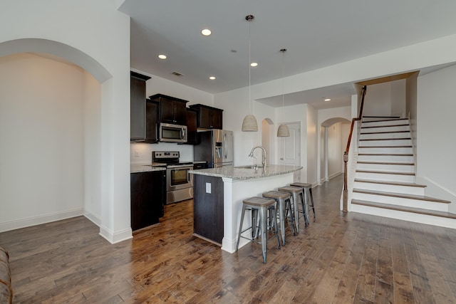 kitchen with a breakfast bar, a kitchen island with sink, hanging light fixtures, appliances with stainless steel finishes, and light stone counters