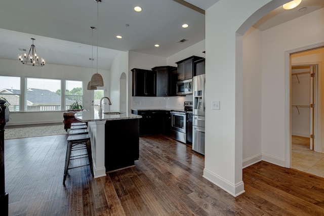 kitchen with light stone countertops, hanging light fixtures, an inviting chandelier, a center island with sink, and appliances with stainless steel finishes