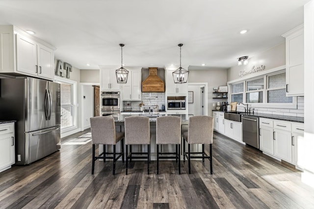 kitchen featuring custom exhaust hood, hanging light fixtures, decorative backsplash, an island with sink, and stainless steel appliances