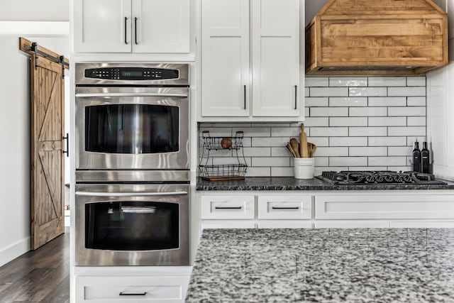 kitchen with stainless steel double oven, a barn door, premium range hood, backsplash, and white cabinets