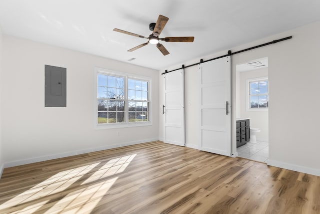 unfurnished bedroom featuring ensuite bathroom, light hardwood / wood-style floors, ceiling fan, electric panel, and a barn door