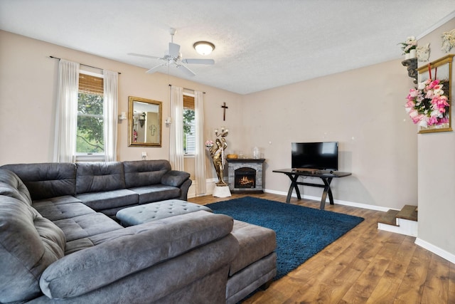 living room featuring ceiling fan, a stone fireplace, wood-type flooring, and a textured ceiling