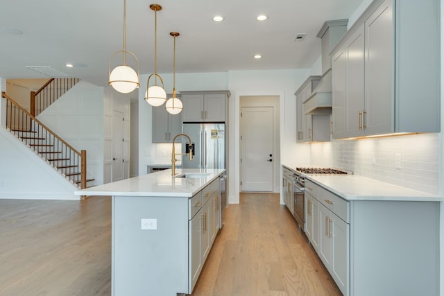 kitchen with stainless steel appliances, tasteful backsplash, light hardwood / wood-style flooring, decorative light fixtures, and a kitchen island with sink