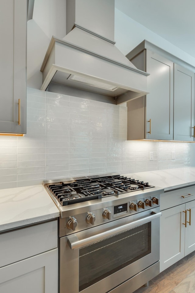 kitchen featuring custom exhaust hood, backsplash, stainless steel stove, light hardwood / wood-style floors, and light stone counters