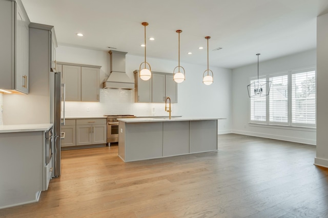 kitchen with stainless steel appliances, an island with sink, decorative light fixtures, gray cabinets, and custom range hood