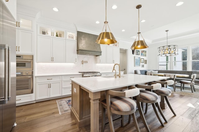 kitchen with pendant lighting, custom exhaust hood, sink, white cabinetry, and a kitchen island with sink