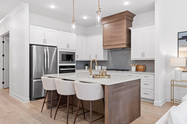 kitchen with custom exhaust hood, white cabinets, hanging light fixtures, an island with sink, and stainless steel appliances