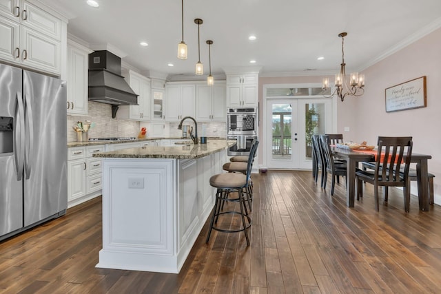 kitchen with white cabinetry, an island with sink, decorative light fixtures, custom range hood, and appliances with stainless steel finishes