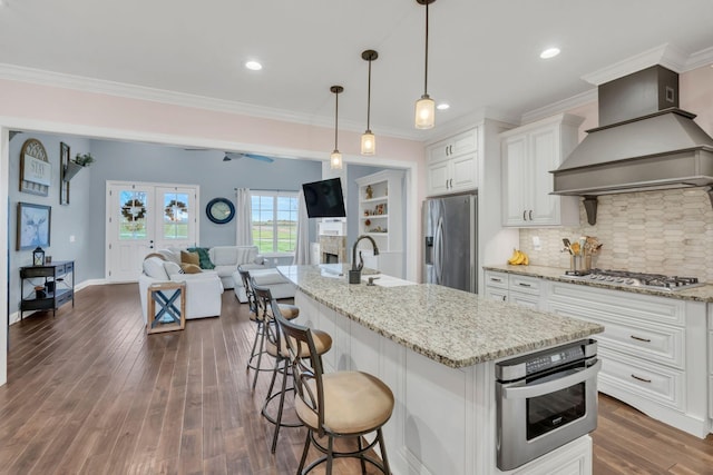 kitchen featuring a kitchen island with sink, hanging light fixtures, custom range hood, appliances with stainless steel finishes, and white cabinetry