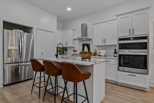 kitchen with stainless steel appliances, wall chimney range hood, a kitchen island with sink, white cabinets, and light wood-type flooring