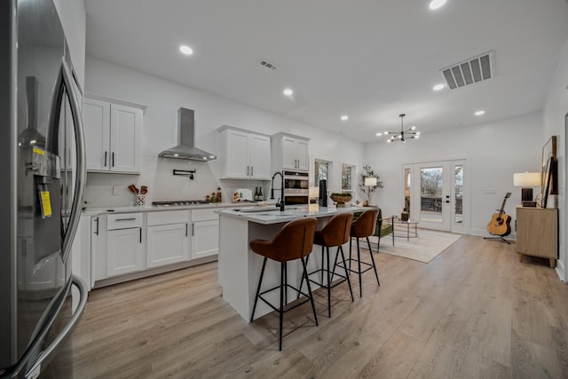 kitchen with appliances with stainless steel finishes, white cabinetry, a kitchen island with sink, and wall chimney range hood