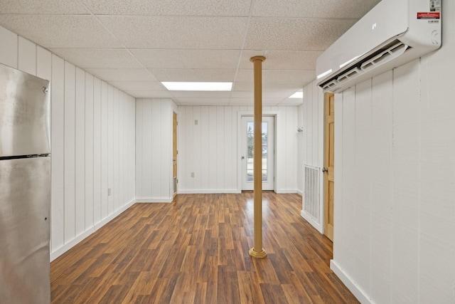 basement with a drop ceiling, an AC wall unit, dark wood-type flooring, and stainless steel refrigerator