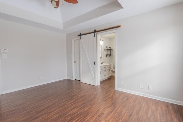 unfurnished bedroom featuring ceiling fan, a raised ceiling, a barn door, dark wood-type flooring, and ensuite bath