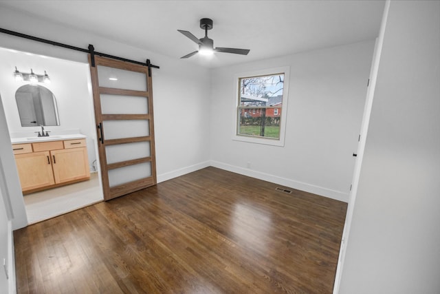 unfurnished bedroom featuring ceiling fan, sink, dark wood-type flooring, a barn door, and ensuite bathroom