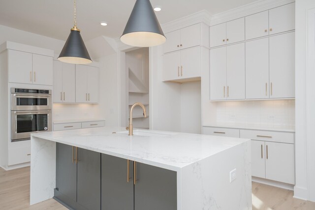 kitchen with stainless steel double oven, decorative light fixtures, white cabinetry, and a kitchen island with sink