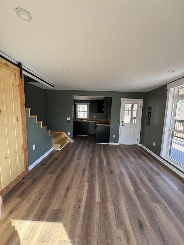 unfurnished living room with stairs, a barn door, baseboards, and dark wood-type flooring