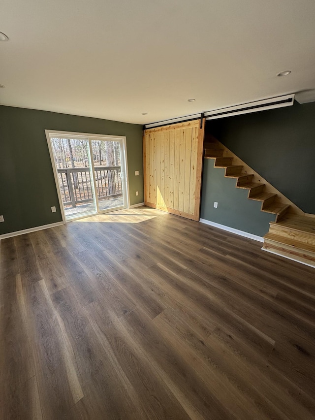 unfurnished living room featuring a barn door, stairway, baseboards, and dark wood-style flooring