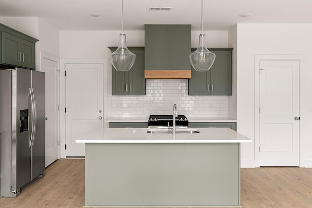 kitchen with premium range hood, hanging light fixtures, stainless steel fridge, and a kitchen island with sink