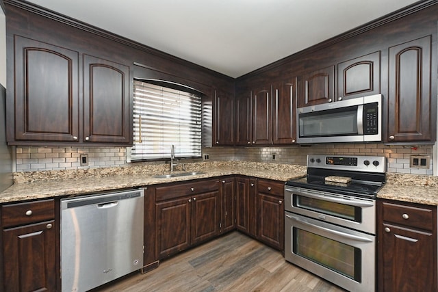 kitchen with appliances with stainless steel finishes, light stone counters, dark brown cabinetry, sink, and light hardwood / wood-style flooring