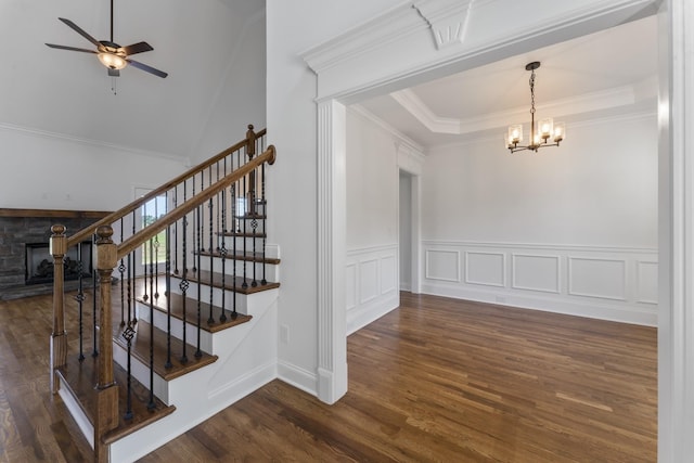 staircase with ornamental molding, ceiling fan with notable chandelier, a tray ceiling, hardwood / wood-style floors, and a stone fireplace