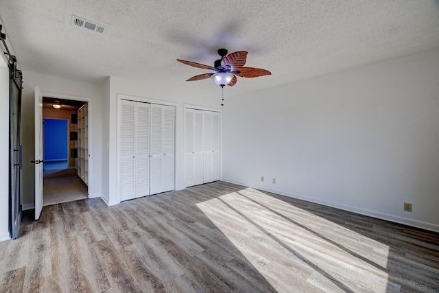 unfurnished bedroom with a textured ceiling, ceiling fan, a barn door, light hardwood / wood-style floors, and multiple closets