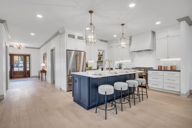 kitchen with hanging light fixtures, premium appliances, premium range hood, a center island with sink, and white cabinets