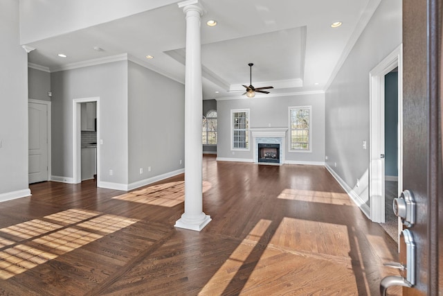 unfurnished living room featuring dark wood-type flooring, ceiling fan, a fireplace, a tray ceiling, and decorative columns