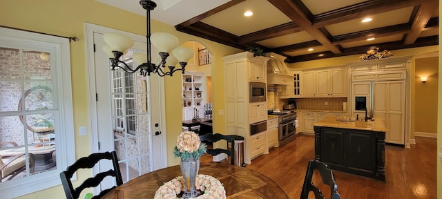 kitchen with coffered ceiling, a center island with sink, appliances with stainless steel finishes, hanging light fixtures, and beam ceiling