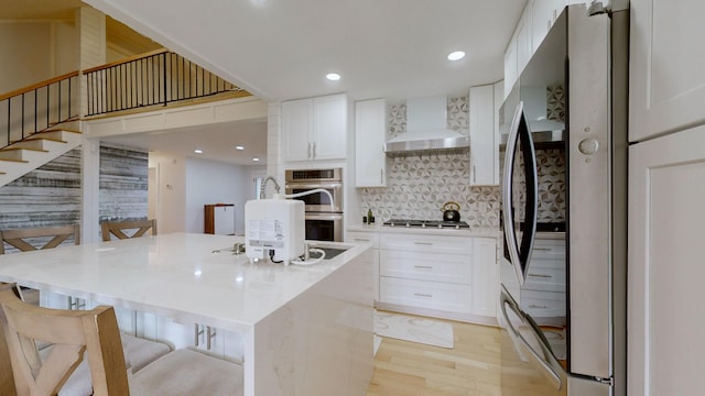 kitchen with a kitchen island with sink, a kitchen breakfast bar, wall chimney range hood, white cabinetry, and stainless steel appliances