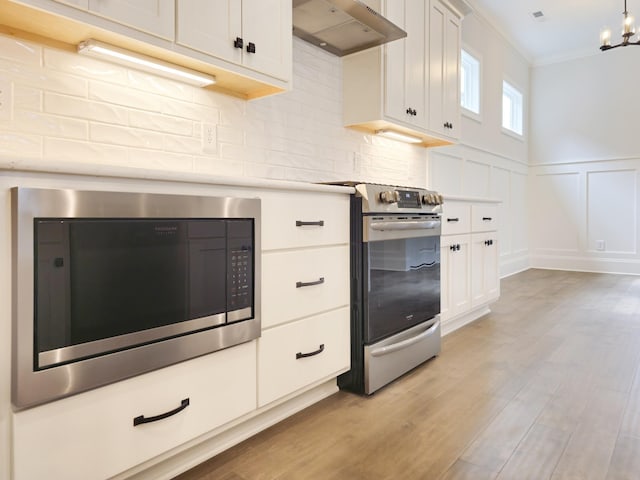 kitchen with exhaust hood, white cabinets, crown molding, stainless steel appliances, and a chandelier