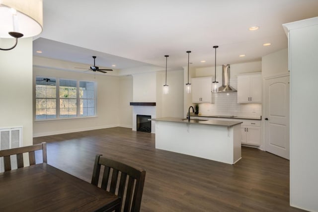 kitchen with sink, wall chimney range hood, decorative light fixtures, a kitchen island with sink, and white cabinets