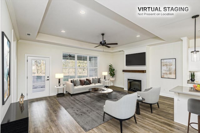 living room featuring ceiling fan, a brick fireplace, hardwood / wood-style floors, a tray ceiling, and ornamental molding