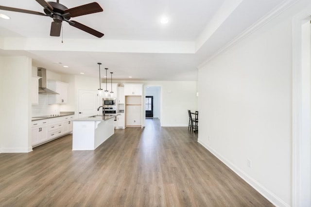 kitchen with appliances with stainless steel finishes, wall chimney range hood, white cabinets, hanging light fixtures, and an island with sink