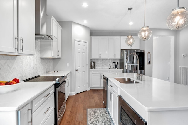 kitchen with appliances with stainless steel finishes, wall chimney exhaust hood, decorative light fixtures, white cabinetry, and an island with sink