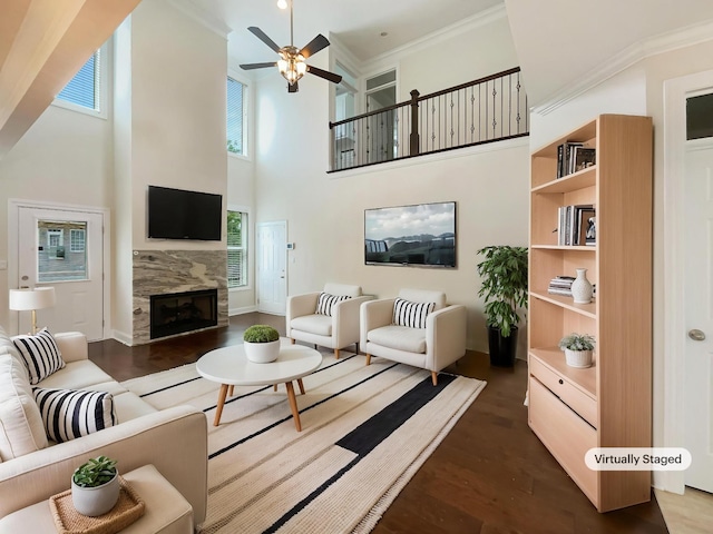 living room featuring a ceiling fan, a tiled fireplace, dark wood-style flooring, a towering ceiling, and ornamental molding