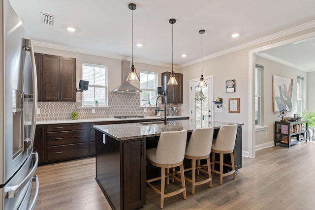 kitchen featuring a center island with sink, appliances with stainless steel finishes, wall chimney range hood, and light wood-type flooring