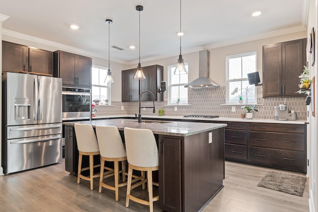 kitchen featuring wall chimney range hood, dark brown cabinets, an island with sink, and appliances with stainless steel finishes