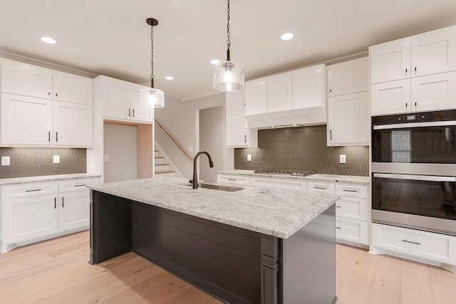 kitchen featuring white cabinetry, sink, multiple ovens, pendant lighting, and a kitchen island with sink