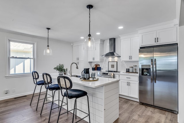 kitchen with a kitchen island with sink, wall chimney range hood, appliances with stainless steel finishes, decorative light fixtures, and white cabinetry