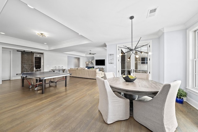 dining room with ornamental molding, ceiling fan with notable chandelier, a raised ceiling, a barn door, and hardwood / wood-style flooring